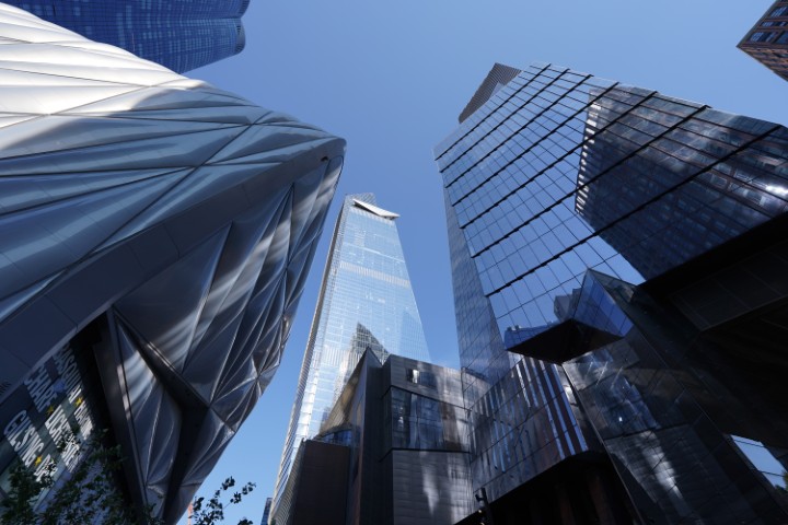 Multiple glass buildings in the financial district of NYC, taken from the ground, looking up. The sky is blue and clear.